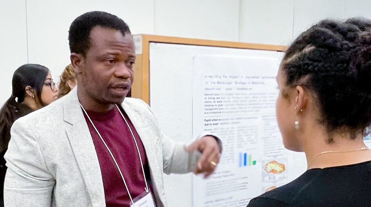 A participant presents research at an academic poster session, gesturing as he speaks with another attendee holding a plate of food. The poster behind them includes text and charts related to urban planning or environmental studies, and other participants are visible in the background.
