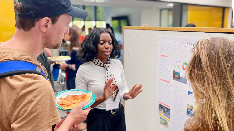 A student presents research at an academic poster session, gesturing as she explains her project to two attendees. One listens while holding a plate of pizza, and another stands beside the display board. The poster features charts and images related to urban development and planning.