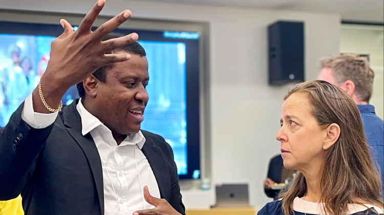 Two people in conversation during an academic event. One gestures expressively while explaining a concept, and the other listens thoughtfully, holding a water bottle. A presentation screen and other attendees are visible in the background.