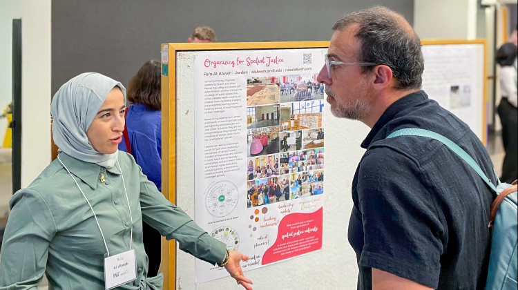 A student presents her research at a poster session, gesturing toward her project while holding a tablet. She speaks with an attendee who listens closely, wearing a backpack. The poster behind them, titled Organizing for Spatial Justice, includes text and photos about community and spatial equity initiatives.