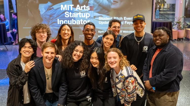 A group of students pose together and smile at the camera during the MIT Arts Startup Incubator event. They stand in front of a large presentation screen displaying event details, conveying a sense of energy, creativity, and collaboration.