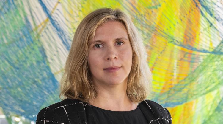 Portrait of MIT Professor Caitlin Mueller standing in front of a colorful abstract background composed of blue, green, and yellow brushstrokes. The person has shoulder-length blonde hair and a calm expression, wearing a dark patterned jacket over a black top.
