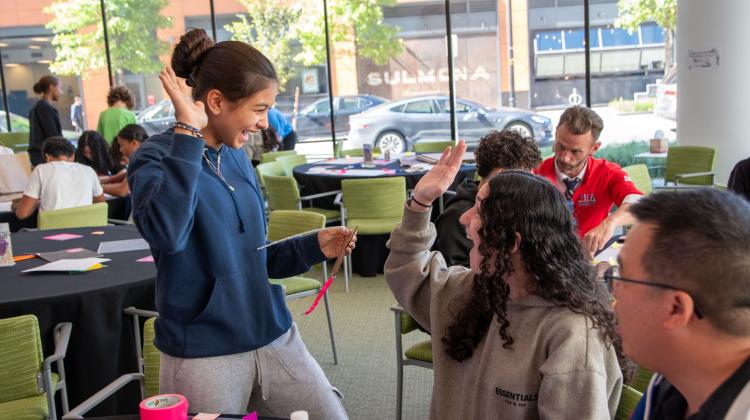 Students collaborate in a bright classroom setting, smiling and giving each other a high five during a creative workshop. Tables around them are covered with colorful materials, paper, and tape, as other participants work in the background near large windows overlooking the street.
