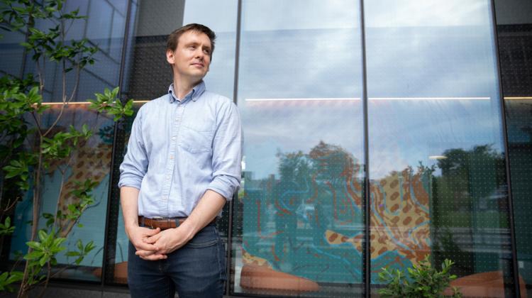 Portrait of Nick Allen standing outdoors in front of a modern glass building. Reflections of trees and sky appear on the windows behind them, along with colorful abstract patterns inside the building. The person wears a light blue shirt and looks slightly to the side with a thoughtful expression.