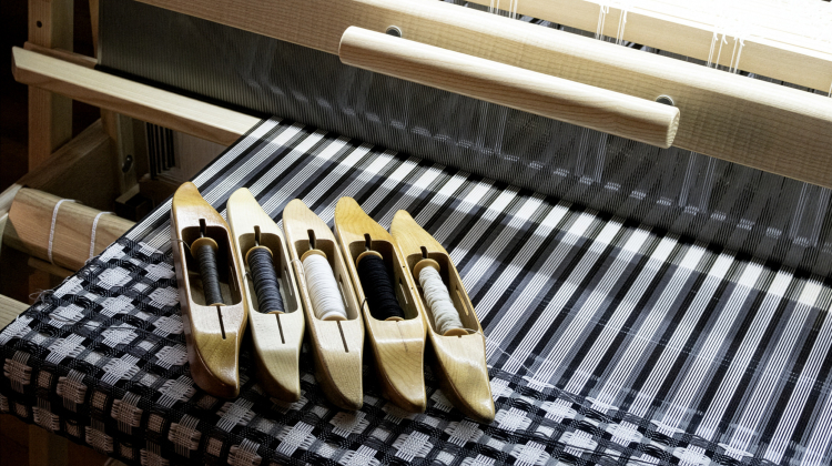 Close-up of a wooden weaving loom with black and white patterned fabric in progress. Several wooden shuttles loaded with dark and light threads rest neatly on the woven surface.