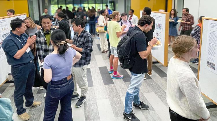 Students and researchers gather in a bright hallway for an academic poster session. Groups of attendees engage in conversation while viewing design and planning projects displayed on large boards. Some hold plates of food as they exchange ideas in a lively, collaborative setting.