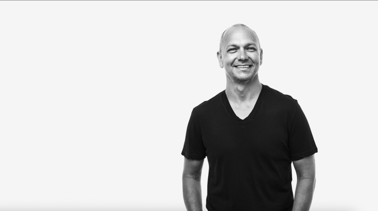 Black-and-white portrait of Tony Fadell standing against a plain light background, smiling and wearing a short-sleeved V-neck T-shirt. The composition is minimal and open, emphasizing a relaxed and approachable demeanor.