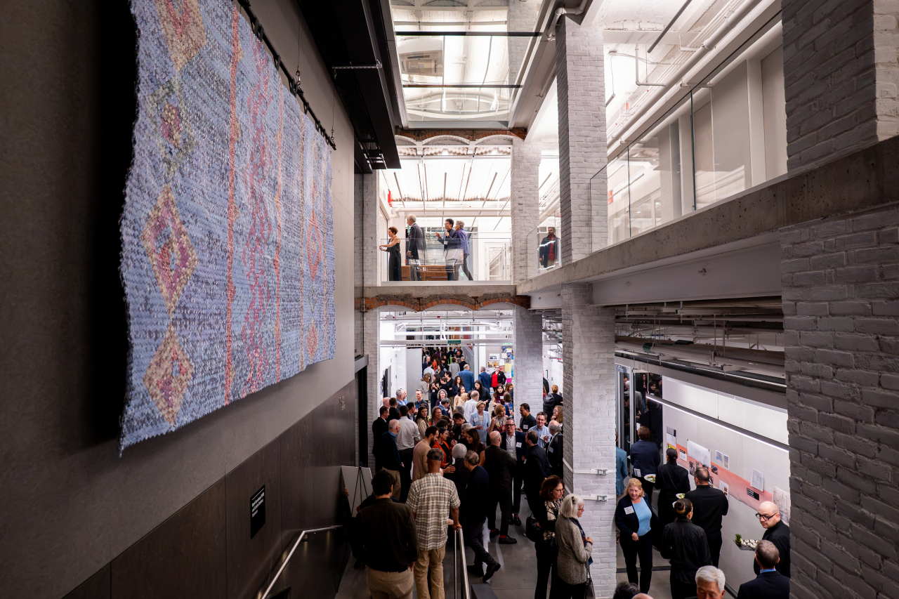 Guests gather in the atrium of MIT’s MET Warehouse during the building’s dedication ceremony, surrounded by historic brick columns, galleries, and a large textile installation.