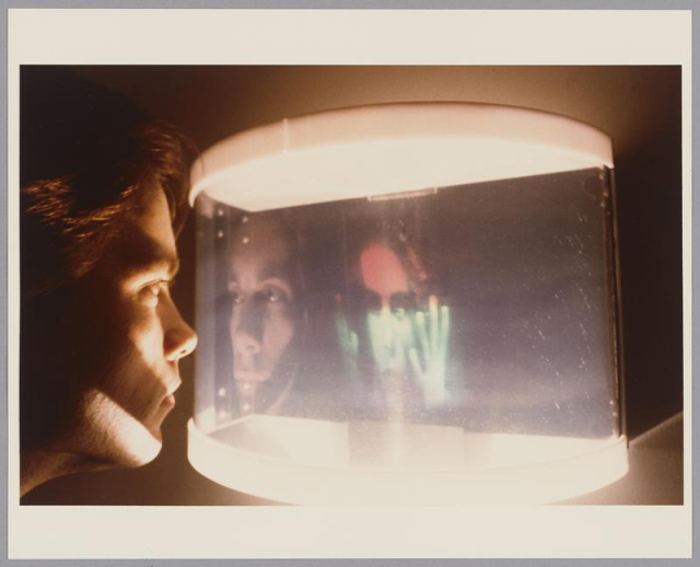 Photo from the Media Lab highlighting a visual arts project. A student stares into a glass screen with a faint projection on the other side.