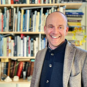 Man with short hair smiling, wearing navy shirt and light brown jacket. Window and bookshelf in the background. 
