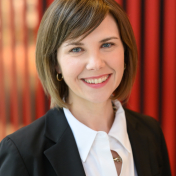 A woman with short hair, smiling, wearing a white shirt and black jacket. Red striped background. 